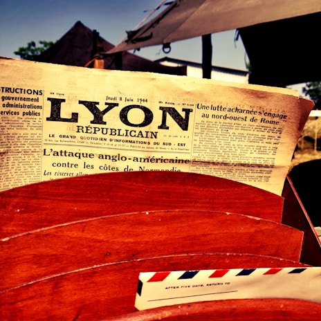 A vintage newspaper with the headline 'Lyon Républicain' is prominently displayed, dated June 8, 1944. The newspaper is placed inside a wooden receptacle alongside an airmail envelope. The background features blurred outdoor elements, possibly a tent or structure.