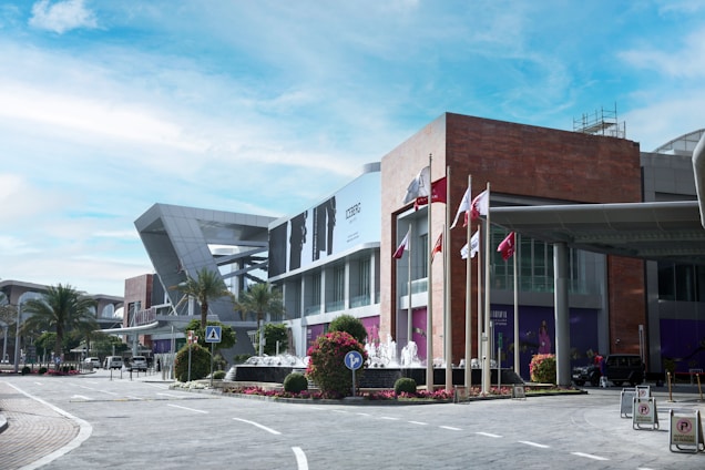 A modern commercial building with angular architecture design and large windows, surrounded by palm trees and manicured flower beds. Several flagpoles with different flags are positioned near the entrance. A road curves in the foreground, with visible traffic signs and a pedestrian crosswalk. The sky appears clear and blue.
