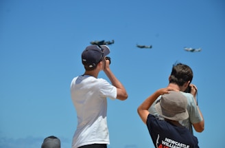 A group of people are looking up at the sky, some with binoculars, as airplanes fly in formation. The scene takes place under a clear blue sky, suggesting an airshow event.