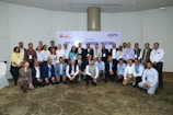 A group of people are gathered in a conference setting, posing for a group photo. Most individuals are dressed in business attire, and many are wearing name badges. Behind them is a banner indicating a meeting or event.