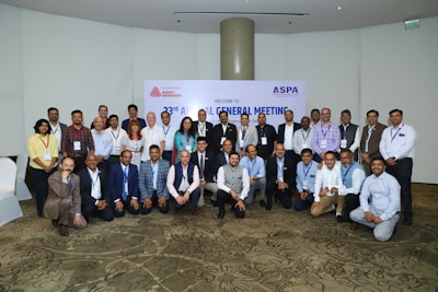 A group of people are gathered in a conference setting, posing for a group photo. Most individuals are dressed in business attire, and many are wearing name badges. Behind them is a banner indicating a meeting or event.