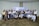 A group of people are gathered in a conference setting, posing for a group photo. Most individuals are dressed in business attire, and many are wearing name badges. Behind them is a banner indicating a meeting or event.
