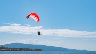 A paraglider soars through the clear blue sky above a body of water. The parachute is a vibrant red with white designs. In the background, a landscape of distant hills and a shoreline is visible, suggesting a coastal location.