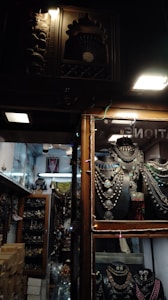 Inside a dimly lit store, rows of intricately crafted jewelry displayed on black holders catch the eye. Necklaces with large pendants and colorful stones are arranged neatly. The shop has wooden frames and showcases with decorative carvings. Shelves in the background are filled with similar ornate jewelry and artifacts.