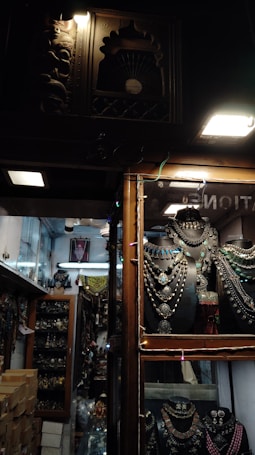 Inside a dimly lit store, rows of intricately crafted jewelry displayed on black holders catch the eye. Necklaces with large pendants and colorful stones are arranged neatly. The shop has wooden frames and showcases with decorative carvings. Shelves in the background are filled with similar ornate jewelry and artifacts.