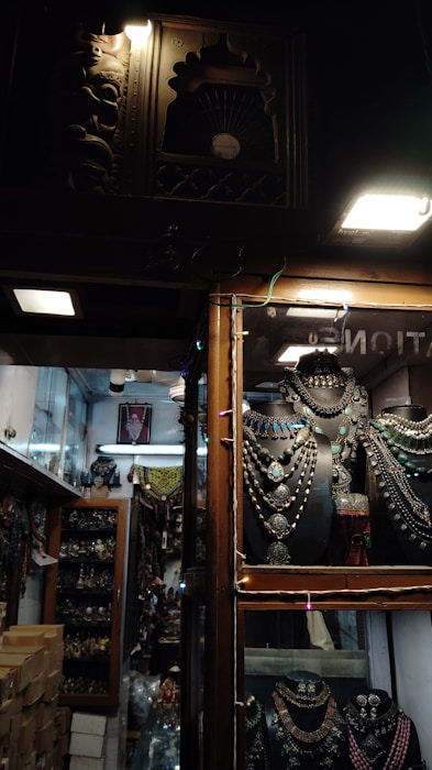 Inside a dimly lit store, rows of intricately crafted jewelry displayed on black holders catch the eye. Necklaces with large pendants and colorful stones are arranged neatly. The shop has wooden frames and showcases with decorative carvings. Shelves in the background are filled with similar ornate jewelry and artifacts.
