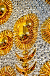 An ornate and intricate design featuring patterns of golden, sun-like shapes and crescents against a background studded with shiny metallic spheres. The design elements reflect light and objects, creating mirrored effects. The reflective materials create a luxurious and opulent appearance.