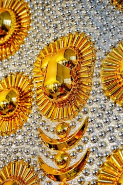An ornate and intricate design featuring patterns of golden, sun-like shapes and crescents against a background studded with shiny metallic spheres. The design elements reflect light and objects, creating mirrored effects. The reflective materials create a luxurious and opulent appearance.