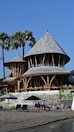 A scenic beachside setting featuring a unique bamboo structure with conical roofs. The building is surrounded by palm trees under a clear blue sky. In the foreground, people relax on beach chairs and under umbrellas on the sandy beach.
