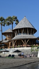 A scenic beachside setting featuring a unique bamboo structure with conical roofs. The building is surrounded by palm trees under a clear blue sky. In the foreground, people relax on beach chairs and under umbrellas on the sandy beach.