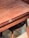 A wooden table with a smooth, polished surface and visible wood grain. It features a brass latch mechanism on the side. There is a wooden chair with a dark seat partially visible underneath the table.