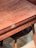 A wooden table with a smooth, polished surface and visible wood grain. It features a brass latch mechanism on the side. There is a wooden chair with a dark seat partially visible underneath the table.