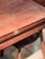 A wooden table with a smooth, polished surface and visible wood grain. It features a brass latch mechanism on the side. There is a wooden chair with a dark seat partially visible underneath the table.