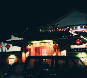 A traditional Chinese building is decorated with red lanterns, illuminated softly against the night sky. The structure features ornamental details and a vehicle is parked in front, partially visible. The scene captures a sense of cultural celebration or gathering.