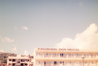 A hotel building with the name 'Panorama-Don Miguel' written on the top is visible under a clear, pale blue sky with some fluffy white clouds. The hotel appears to have multiple floors with balconies. Additional smaller buildings can be seen in the background along with a lamppost in the foreground.