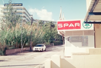 A street corner featuring a SPAR store with a prominent sign. A white car is parked along the road, which is lined with tall grasses and trees. In the background, a modern building with balconies can be seen under a partly cloudy sky.