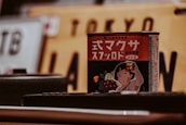 A product with Japanese text on a red can is prominently featured. The can displays an illustration of a child holding up a toast with jam, surrounded by images of fruits such as lemon and grapes. In the background, a blurred Japanese license plate with the word 'Tokyo' is visible.
