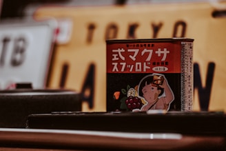 A product with Japanese text on a red can is prominently featured. The can displays an illustration of a child holding up a toast with jam, surrounded by images of fruits such as lemon and grapes. In the background, a blurred Japanese license plate with the word 'Tokyo' is visible.