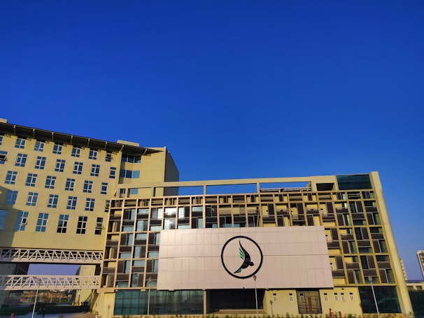 A modern, multi-story building with a contemporary architectural design featuring large windows and a beige facade. A single prominent logo is displayed at the center of the building’s front side. The sky is clear and vividly blue, providing a stark contrast to the building.