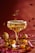 A glamorous cocktail glass filled with a golden liquid, garnished with sugar around the rim. Gold and red festive decorations like glittery ornaments and small golden tree confetti are scattered around the table, conveying a holiday or celebratory theme.