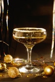 A crystal-clear cocktail glass filled with a golden liquid, featuring a sugar rim. Surrounding the glass are shimmering gold ornaments and glittery decorations, set against a dark background with subtle reflective surfaces.