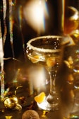 A festive drink with a sugared rim is in focus, surrounded by golden and glittery decorations. The drink contains pieces of fruit, possibly orange slices, and is in a champagne coupe glass. There are colorful light reflections and tinsel adding to the festive atmosphere.