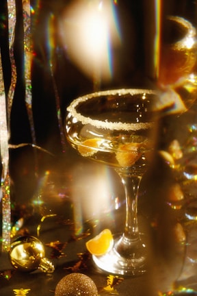 A festive drink with a sugared rim is in focus, surrounded by golden and glittery decorations. The drink contains pieces of fruit, possibly orange slices, and is in a champagne coupe glass. There are colorful light reflections and tinsel adding to the festive atmosphere.
