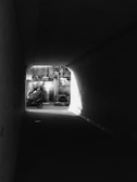 A black and white photograph captures a motorcycle exiting a dimly lit tunnel into a brighter outdoor space. On the motorcycle are two riders wearing helmets. The tunnel is dark, contrasting sharply with the light shining in from outside, highlighting the vehicle and riders.