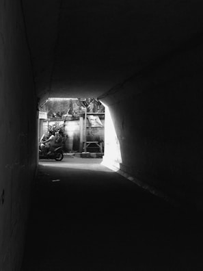 A black and white photograph captures a motorcycle exiting a dimly lit tunnel into a brighter outdoor space. On the motorcycle are two riders wearing helmets. The tunnel is dark, contrasting sharply with the light shining in from outside, highlighting the vehicle and riders.
