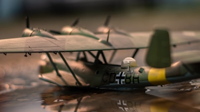 A detailed view of a model seaplane featuring military insignia and camouflage coloring is displayed. The aircraft is positioned on a reflective surface, suggesting a water setting. The focus highlights intricate details such as the cockpit and markings.