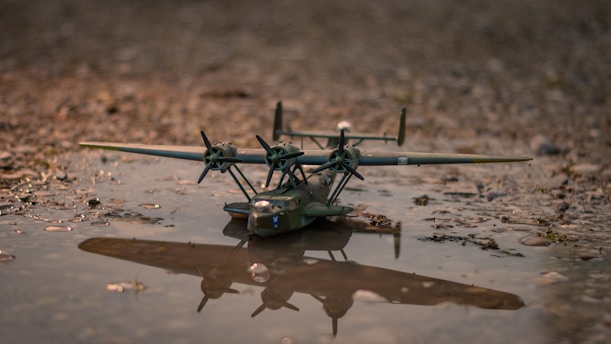 A small model airplane rests in a shallow puddle on a rocky or gravelly surface. The airplane is detailed with multiple propellers and its reflection is visible in the water. The scene appears calm and is captured in natural light, giving it a serene feeling.