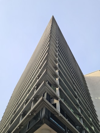 A modern, triangular-shaped building that features a series of long, horizontal panels. The perspective is from the ground looking up, highlighting the sharp angles and geometric design against a clear blue sky.