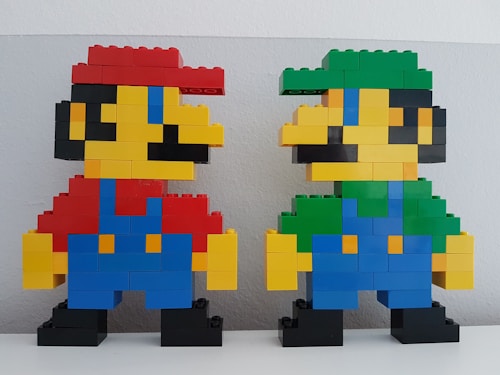 Two pixelated figures made from LEGO bricks resembling video game characters. The figure on the left is constructed with predominantly red, blue, yellow, and black bricks, featuring a red hat and blue overalls. The figure on the right is similarly constructed with green, blue, yellow, and black bricks, featuring a green hat and blue overalls.