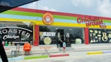 A colorful restaurant exterior with a sign reading 'Chicken Broast'. The building has a bright red, yellow, and green trim. Large windows display images of food like burgers and a salad, along with text such as 'Eat Healthy' and 'Roasted Chicken'. In front, there are several chairs and a small table, and a child is standing near the glass door entrance.