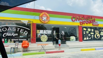 A colorful restaurant exterior with a sign reading 'Chicken Broast'. The building has a bright red, yellow, and green trim. Large windows display images of food like burgers and a salad, along with text such as 'Eat Healthy' and 'Roasted Chicken'. In front, there are several chairs and a small table, and a child is standing near the glass door entrance.