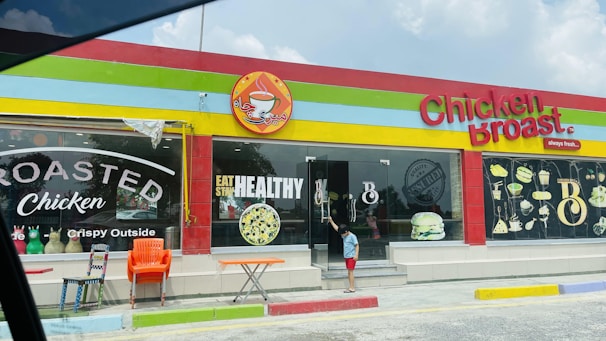A colorful restaurant exterior with a sign reading 'Chicken Broast'. The building has a bright red, yellow, and green trim. Large windows display images of food like burgers and a salad, along with text such as 'Eat Healthy' and 'Roasted Chicken'. In front, there are several chairs and a small table, and a child is standing near the glass door entrance.