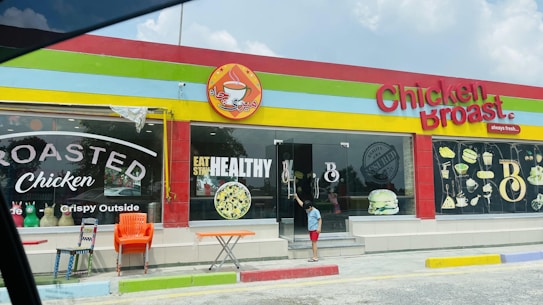 A colorful restaurant exterior with a sign reading 'Chicken Broast'. The building has a bright red, yellow, and green trim. Large windows display images of food like burgers and a salad, along with text such as 'Eat Healthy' and 'Roasted Chicken'. In front, there are several chairs and a small table, and a child is standing near the glass door entrance.
