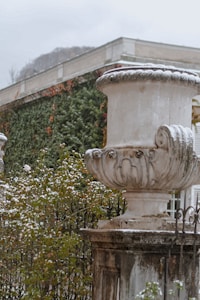 A large, ornate stone urn is set atop a pedestal, covered with a light dusting of snow. Surrounding it are green bushes with scattered red and white flowers. In the background, a building wall is partially covered with climbing ivy, some of which is tinged with autumnal colors.