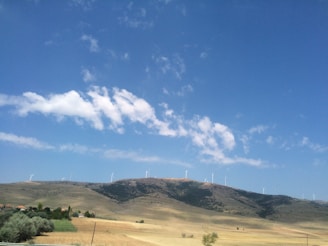 A landscape featuring a range of hills topped with wind turbines under a vast blue sky. A few white clouds are scattered across the sky, while the landscape below is composed of dry, golden fields interspersed with patches of green vegetation and a small cluster of houses.