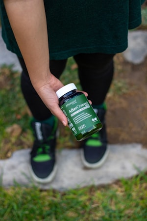 A person is holding a bottle of supplements labeled 'After C Care' with a green and white label. The person is standing on a stone path surrounded by grass, wearing black and green sneakers with black pants and a green top.