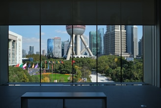 A panoramic view from inside a building featuring large windows overlooking a vibrant urban landscape. Prominently displayed is a unique spherical structure with supporting pillars, surrounded by modern skyscrapers. Below, a park area is visible with numerous flagpoles displaying colorful flags.