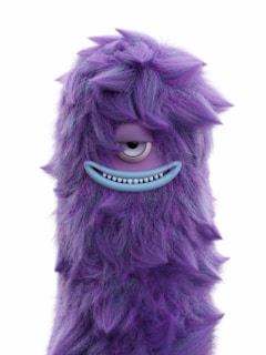 A friendly purple monster with big round eyes and a goofy grin, waving hello in a bright playroom.