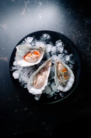 Three opened oysters are placed on a bed of crushed ice atop a dark plate. The oysters have a glossy appearance with contrasting orange adornments, possibly sauce or garnishes, highlighting their rich and fresh texture.