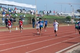 A group of young athletes sprinting on a running track, competing in a race with numbered bibs. Spectators are visible in the background under a sheltered area, observing the event. The sky is clear, contributing to a bright, energetic atmosphere.