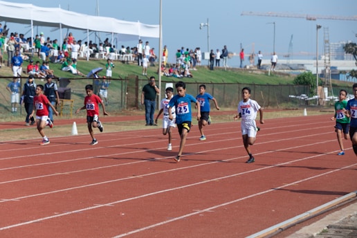 A group of young athletes sprinting on a running track, competing in a race with numbered bibs. Spectators are visible in the background under a sheltered area, observing the event. The sky is clear, contributing to a bright, energetic atmosphere.