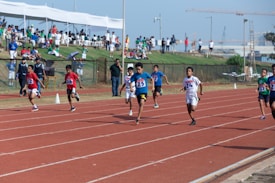 A group of young athletes sprinting on a running track, competing in a race with numbered bibs. Spectators are visible in the background under a sheltered area, observing the event. The sky is clear, contributing to a bright, energetic atmosphere.