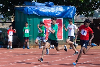 A group of young boys are participating in a sprint race on an outdoor track. They are wearing numbered bibs and athletic attire. In the foreground, several runners are captured mid-stride, conveying a sense of speed and competition. A green container and a tree are visible in the background, providing shade.