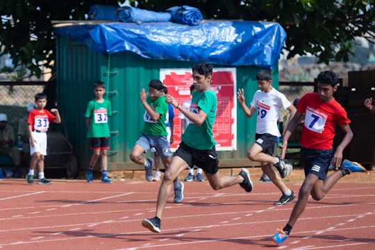 A group of young boys are participating in a sprint race on an outdoor track. They are wearing numbered bibs and athletic attire. In the foreground, several runners are captured mid-stride, conveying a sense of speed and competition. A green container and a tree are visible in the background, providing shade.