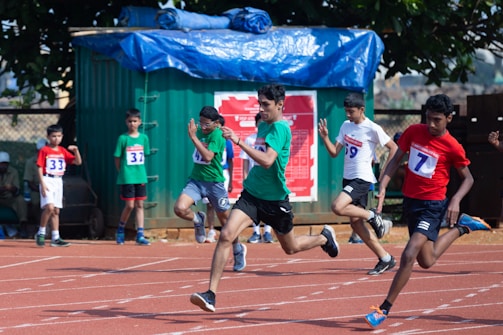 A group of young boys are participating in a sprint race on an outdoor track. They are wearing numbered bibs and athletic attire. In the foreground, several runners are captured mid-stride, conveying a sense of speed and competition. A green container and a tree are visible in the background, providing shade.