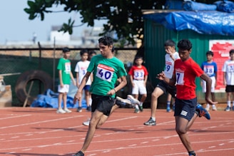 Several young athletes are sprinting on a track in what appears to be a race, with spectators and other participants visible in the background. The runners are wearing numbered bibs and colorful sports attire, competing intensely under clear skies.